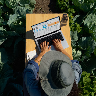 Vue en plongée d’une femme coiffée d’un chapeau à larges bords et installée à une table de jardin, préparant une présentation d’affaires sur un MacBook.