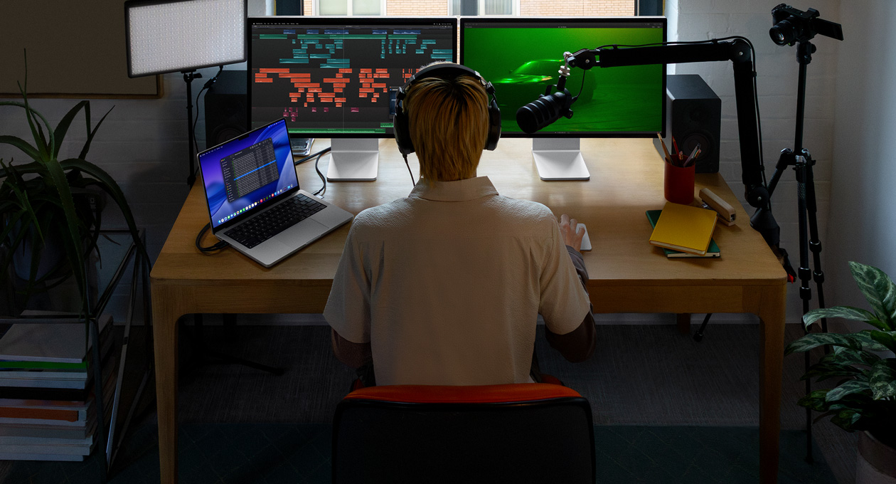 Person sitting at desk with a mic and headphones in front of three devices, Mac Studio Display screen, audio editing tools, sound clips in different blocks of colour, a second Mac Studio Display screen showing, moody shot of car in fog with green lighting, MacBook Pro 16 screen showing Finder app on desktop showing Files for Final Cut Pro, dock, blue stylised desktop background