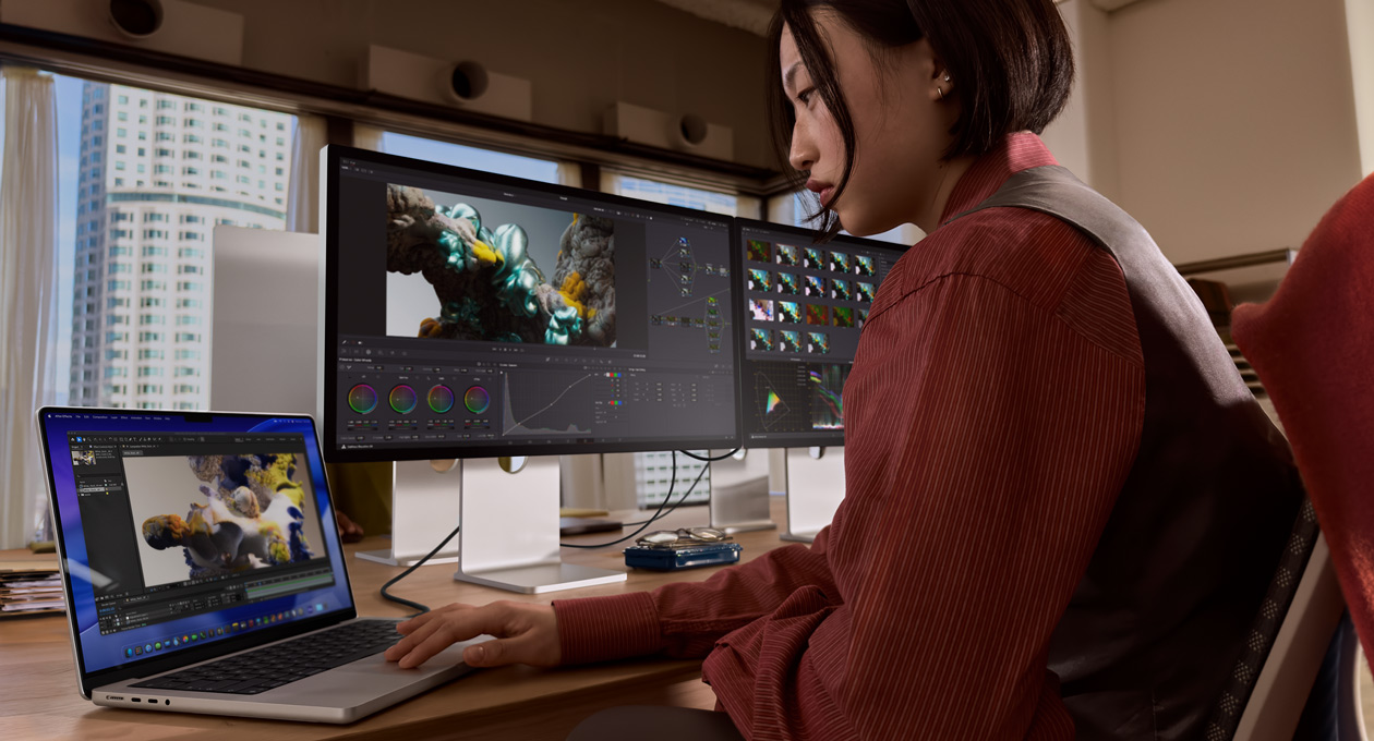 A person in an office setting works on a MacBook laptop that is connected to Studio Display XDR by a Thunderbolt 5 port
