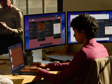 Un equipo de empleados sentados alrededor de una mesa en una oficina. Una mujer sentada en su escritorio trabaja en un MacBook con varios monitores. Sus colegas están reunidos conversando.