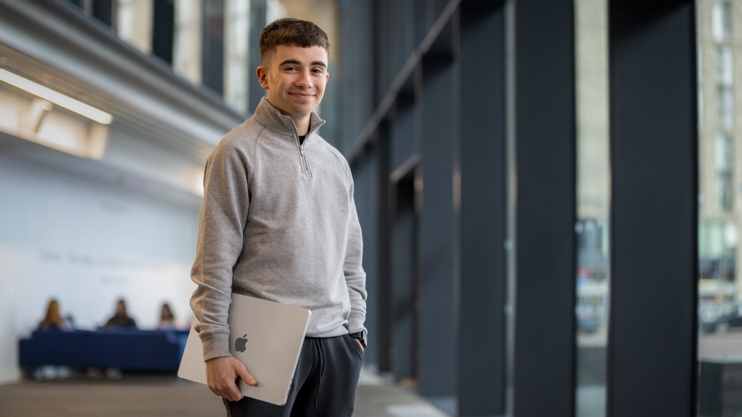 En studerende står på gangen i en moderne bygning og holder en bærbar MacBook, mens han smiler til kameraet
