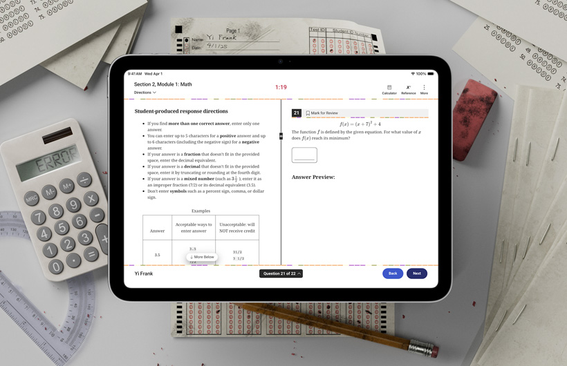 En iPad ligger på et bord blandt fysiske prøver og skoleredskaber. På skærmen vises en vejledning i at gennemføre matematikdelen af en standardiseret prøve.
