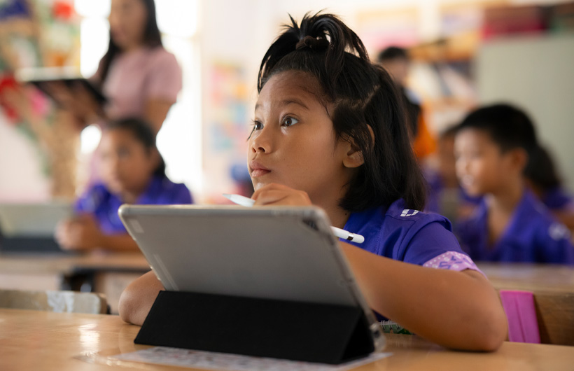 Una bambina seduta al banco guarda verso la cattedra, seguendo con grande attenzione. Usa un iPad e tiene in mano un’Apple Pencil.