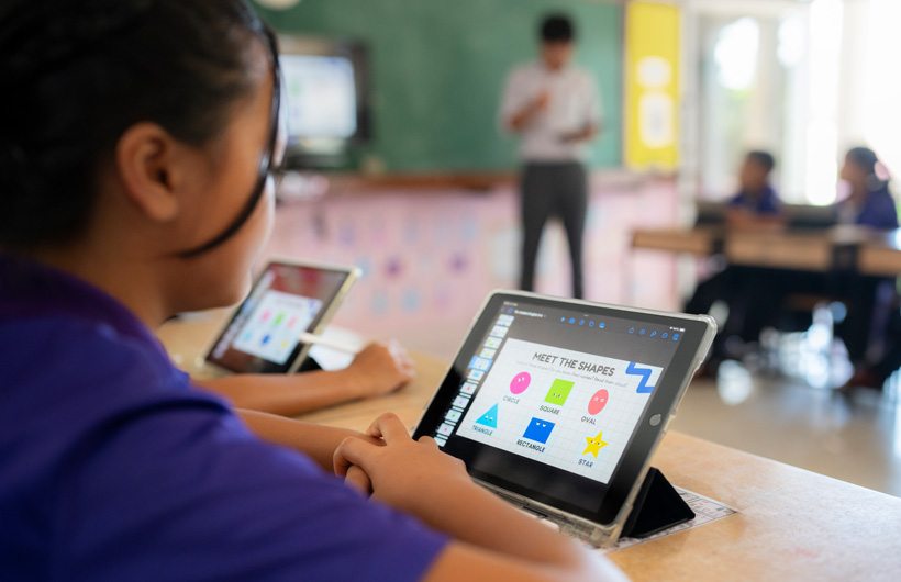 Primo piano di una studentessa seduta al banco che usa un iPad. Sullo schermo ci sono forme colorate con il titolo “Scopri le forme”.