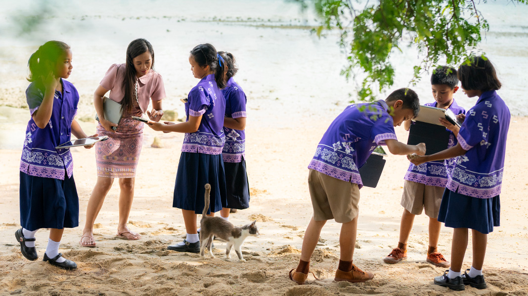 Sei studenti della scuola primaria in uniformi viola, in piedi su una spiaggia sabbiosa con in mano gli iPad. L’insegnante sta aiutando una studentessa su un iPad e c’è un gatto che cammina in mezzo al gruppo.