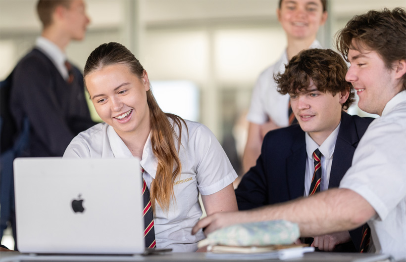 En gruppe elever i skoleuniformer samlet omkring en MacBook smiler og samarbejder.