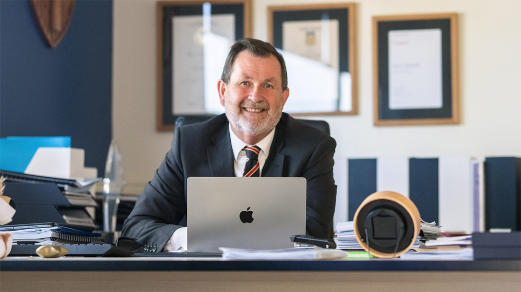 En rektor sidder ved sit skrivebord bag en åben bærbar Apple-computer og smiler til kameraet.