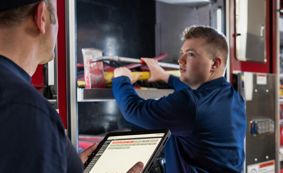 Dois membros da equipa inspecionam ferramentas e equipamento num camião de bombeiros; um segura um iPad enquanto o outro faz a inspeção