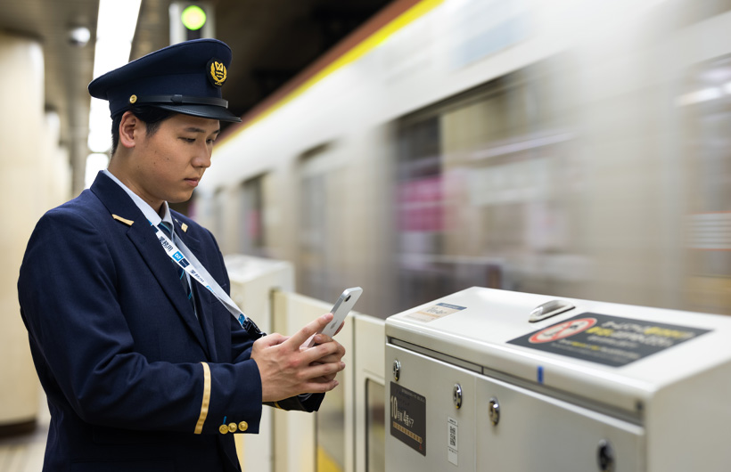 A station attendant stands on the platform and uses his iPhone to scan a QR code while a train passes through.