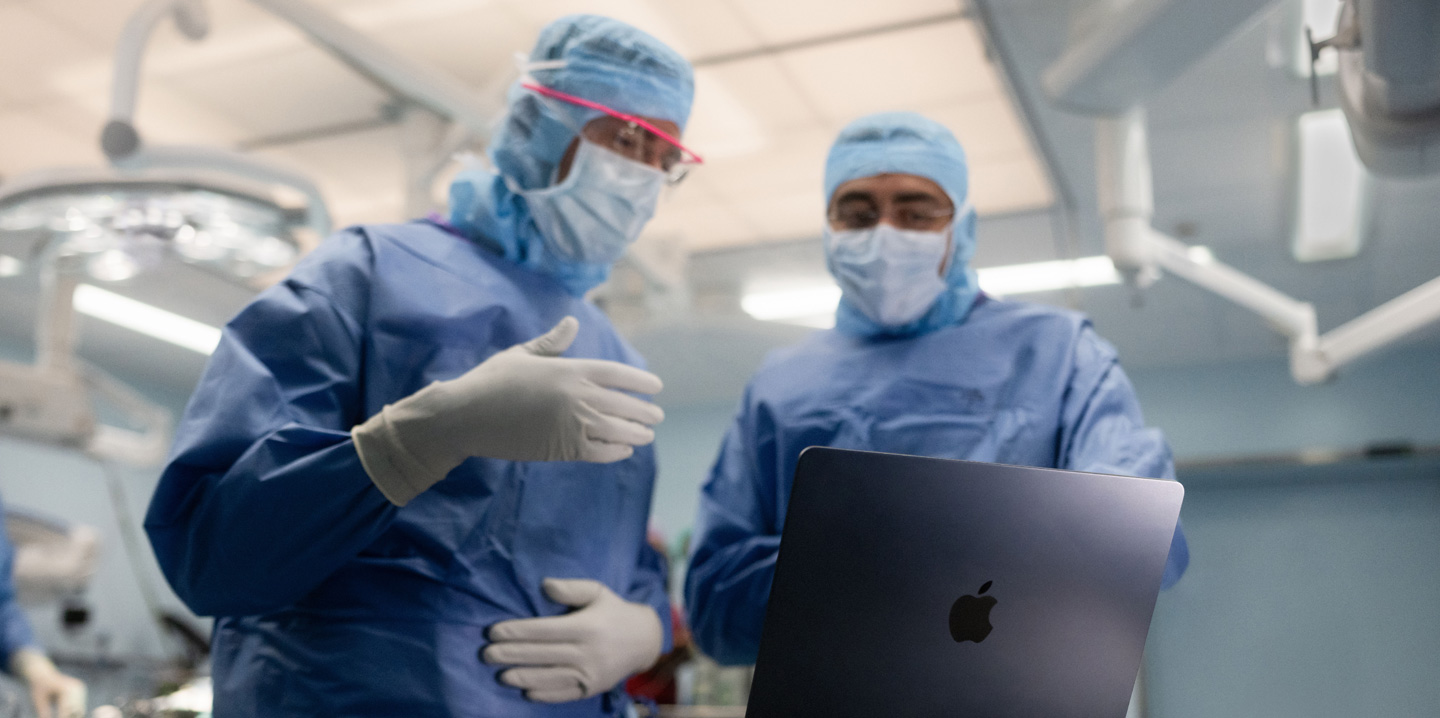 Two surgeons in the operating room look at a MacBook, and one gestures with their hand while the other observes