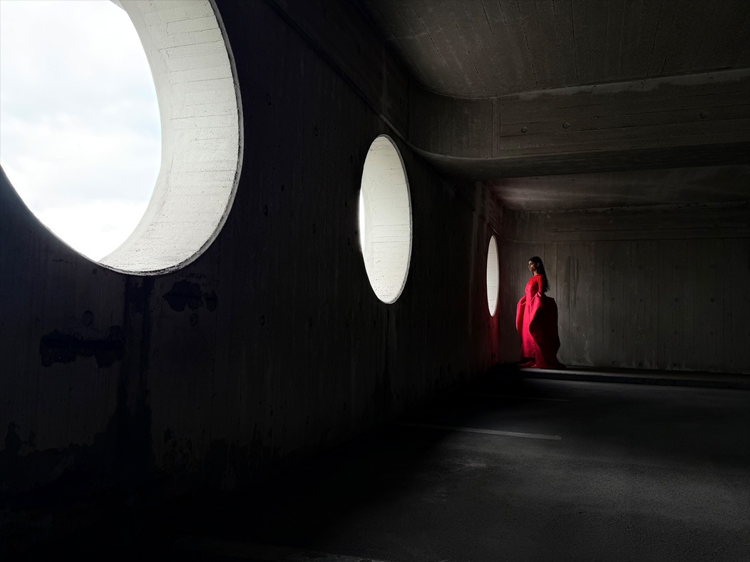 A woman wearing a long red dress stands in an empty room being photographed. The Photo app toggles through the following focal lengths to demonstrate the capabilities of the pro camera system: 200 mm, 100 mm, 48 mm, 35 mm, 28 mm, 13 mm, and macro